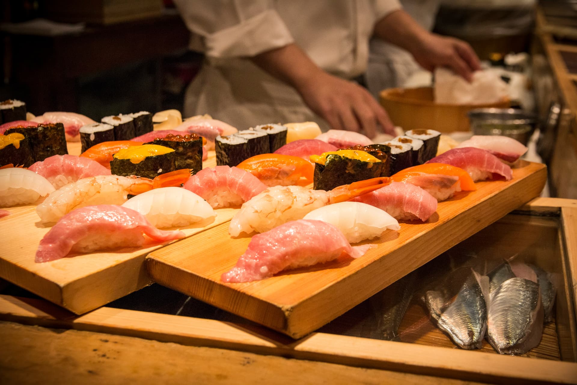 surtito de sushi de pescado fresco donde se ve salmon, atun y otros pescados con el cocinero preparándolo de fondo. Sushi sobre tabla de madera.